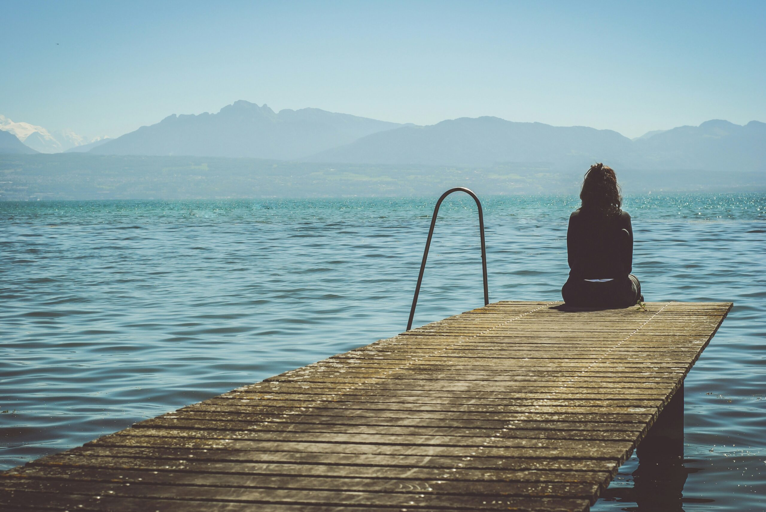Image of a woman's back sitting on the end of a pier. The image has a lonely feel to it.